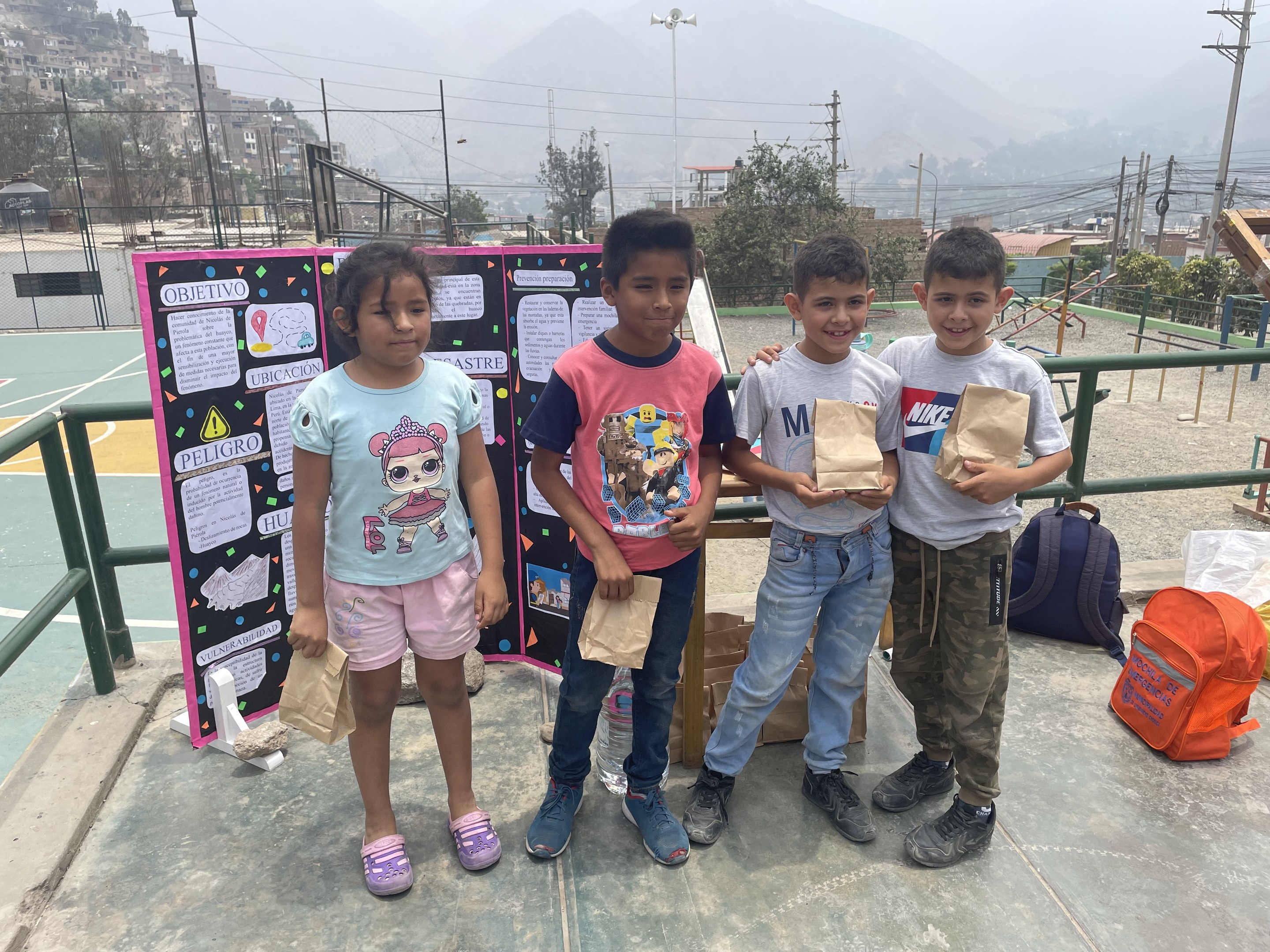 Gruppe von Kindern, die Papiertüten auf einem Basketballfeld halten, mit Tüten und Gegenständen auf dem Boden, einer Tafel mit Text links und einem Zaun, Pfosten, Straßenlaternen, Bäumen, Gebäuden, Hügeln und Himmel im Hintergrund.