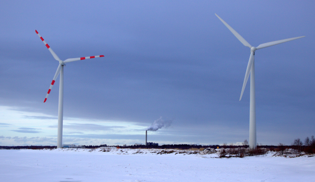 Drei Windkraftanlagen in einem verschneiten Feld, umgeben von Bäumen und Pflanzen, mit Rauch, der aus ihnen aufsteigt, unter einem bewölktem Himmel.