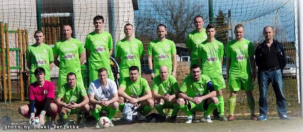 Eine Gruppe von Männern in grünen T-Shirts steht oder hockt auf einem Fußballfeld in der Nähe eines Torpfostens, mit einem Ball davor und verschiedenen Hintergrundelementen wie Bäume, Gebäude und Fahrzeuge.