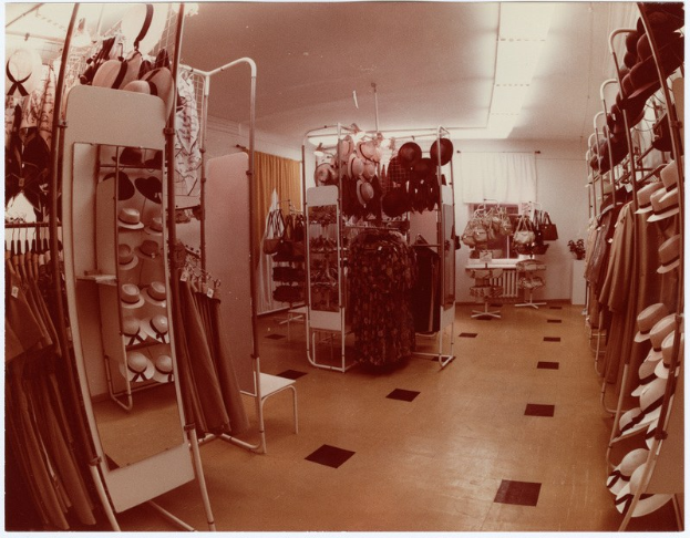 Altes Foto eines Kleiderladens mit Kleiderstangen, Hutständern, einem Spiegel, einem Stuhl, einem Tisch mit Gegenständen, Vorhängen und Deckenbeleuchtung.