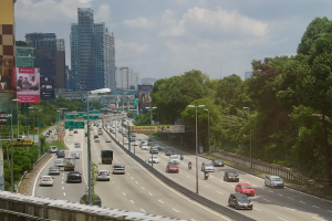 Eine vielbefahrene Autobahn mit zahlreichen Fahrzeugen, gesäumt von hohen Gebäuden, Straßenschildern, Straßenlaternen, Werbetafeln und Bäumen, unter einem bewölkten Himmel.