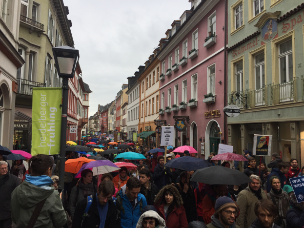 Eine große Gruppe von Menschen mit Schirmen auf der Straße, einige tragen Taschen und halten Schilder mit Text, während sie an einer Klimademonstration in Deutschland teilnehmen.
