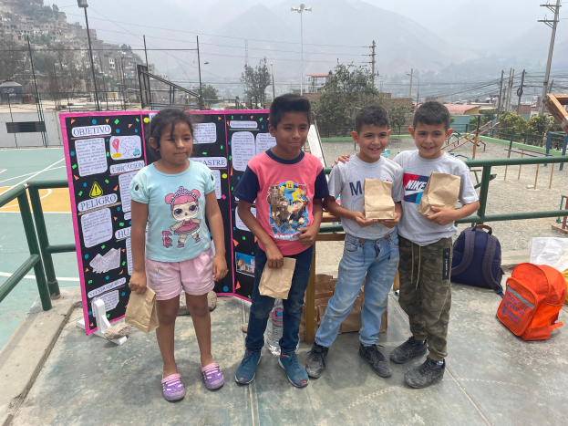 Eine Gruppe von Kindern steht auf einem Basketballfeld, jedes hält einen Papiertüte in der Hand, mit Tüten und Gegenständen rechts daneben und einer Tafel mit Text links daneben, mit Zaun, Pfosten, Straßenlaternen, Strommasten mit Drähten, Bäumen, Gebäuden, Hügeln und Himmel im Hintergrund.
