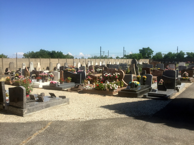 Ein Friedhof mit zahlreichen von Blumen geschmückten Grabsteinen, umgeben von einer Mauer, Strommasten mit Drähten, Bäumen und einem bewölkten Himmel.