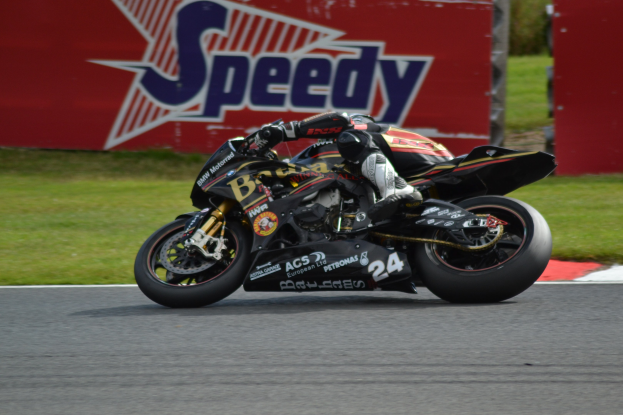 Eine Person in Helm und Schutzkleidung fährt ein Motorrad auf einer Rennstrecke, im Hintergrund ist Gras und eine Tafel mit Text zu sehen.