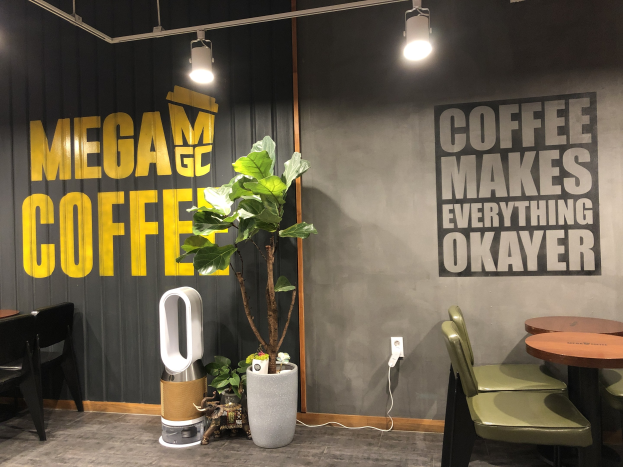 Kaffeehaus-Interieur mit Tischen, Stühlen, einem "Mega Coffee"-Schild an der Wand, einem Topf mit einer Pflanze, einer Bodenstehmaschine und Deckenleuchten.