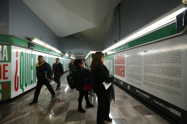 Menschen gehen durch eine hell erleuchtete U-Bahn-Station mit textbedeckten Wänden, einige tragen Taschen.