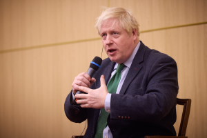 Ein Mann im Anzug und Krawatte sitzt in einem Stuhl und hält ein Mikrofon, sieht aus wie in einer hitzigen Diskussion.