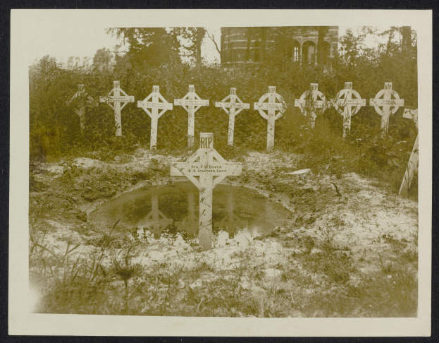 Schwarzes und weißes Foto eines Friedhofs mit zahlreichen Kreuzen im Vordergrund, umgeben von Bäumen und einem Gebäude im Hintergrund, mit einem Gewässer in der Mitte.