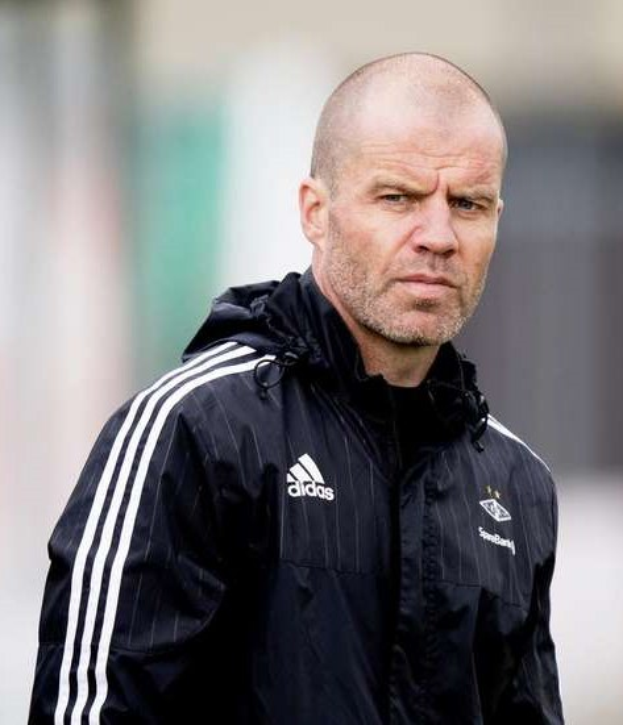 Bald coach in einem blauen Jackett mit weißen Streifen und weißem Kragen, der auf einem Fußballfeld steht und die Hände gefaltet hat