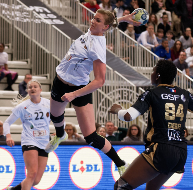 Eine Gruppe von Frauen, die Handball auf einem Platz spielen, mit einem Ball in der Luft, Zuschauern auf Treppen und Geländern im Hintergrund und einem Schild mit der Aufschrift "Handball-Weltmeisterschaft 2019 - Tag 2".