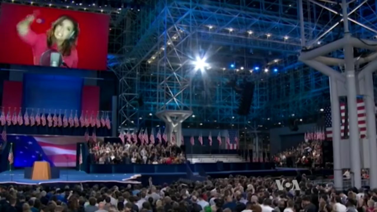 Eine große Zuschauermenge sitzt vor einer Bühne mit einem Podium, Mikrofon, Flaggen, einem Bildschirm mit dem Bild einer Frau und Scheinwerfern und Stangen, was auf eine Präsidentschaftsdebatte hindeutet.