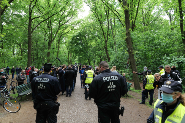 Polizeibeamte vor einer Menge bei einer Outdoor-Veranstaltung, mit Fahrrädern und einer Bank im Vordergrund und Bäumen im Hintergrund.