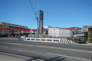 Tankstelle an einer Straßenecke mit parkenden Autos, umgeben von Gebäuden, Strommasten, Schildern, Geländern und Pflanzen unter einem bewölkten Himmel.