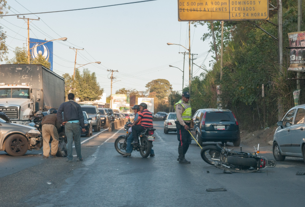 Gruppe von Menschen um ein verunglücktes Motorrad am Straßenrand mit mehreren Fahrzeugen, darunter ein Lastwagen, im Hintergrund und umgebender Infrastruktur.