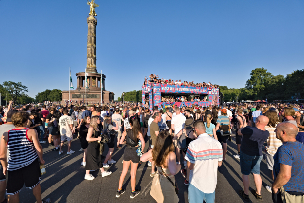 Eine große Menschenmenge steht vor einem Denkmal in Berlin, Deutschland, mit Fahnen, einem Gebäude mit Säulen und einer Statue, Bäumen und einem klaren blauen Himmel im Hintergrund.