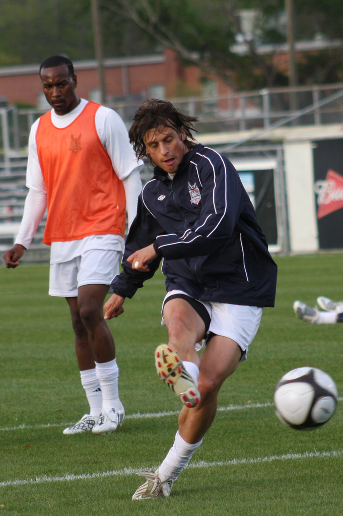 Ein Fußballspieler in einer schwarzen Jacke und weißen Shorts kickt einen Ball, während ein anderer Spieler in einer orangen Jacke hinter ihm steht und ein eingezäunter Bereich mit Bäumen im Hintergrund zu sehen ist.