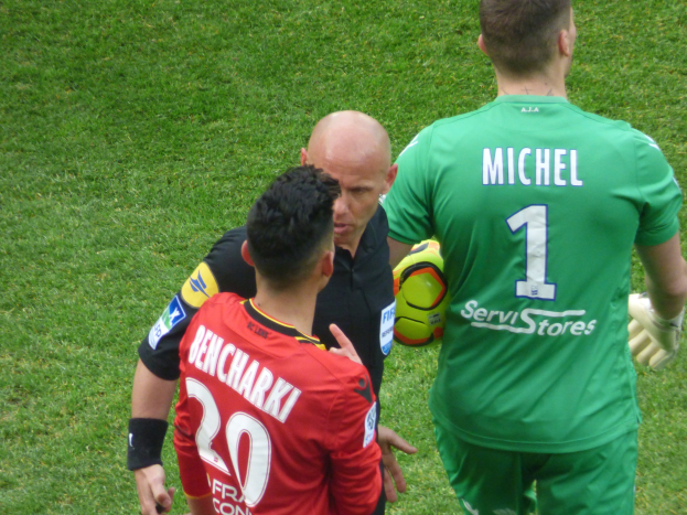 Ein Schiedsrichter spricht mit zwei Fußballspielern auf einem Rasenfeld, wobei einer der Spieler einen Ball hält.