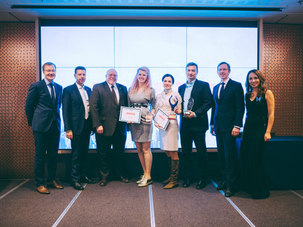 Eine Gruppe strahlender Menschen hält Zertifikate in der Hand und steht vor einem Bildschirm, rechts daneben ein auf schwarzem Tuch gedeckter Tisch und ein Objekt an der Wand im Hintergrund, feiert ihren Sieg bei den International Business Awards 2018.