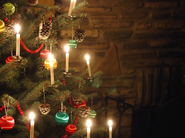 Ein Weihnachtsbaum mit angezündeten Kerzen auf der linken Seite, mit einer Wand im Hintergrund.