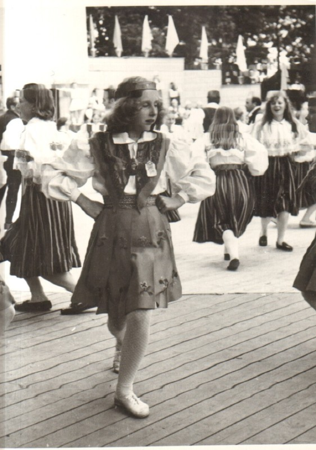 Eine Gruppe von Frauen in traditioneller bayrischer Tracht tanzt auf einem Holzboden, mit einer Wand, Bäumen und anderen Gegenständen im Hintergrund und Text am unteren Rand des Bildes.