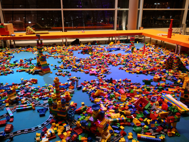 Ein großer Raum mit farbigen Lego-Bauklötzen, die auf dem Boden verstreut sind, umgeben von Tischen und Säulen, mit Glasfenstern im Hintergrund, die Menschen und Lichter zeigen.