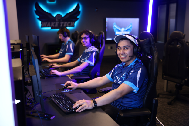 Gruppe von Menschen, die an einem Schreibtisch mit Computern sitzen, Kopfhörer tragen und lächeln, vor Monitoren, Tastaturen und anderen Gegenständen, mit einem Logo und Text an der Wand dahinter und Stühlen rechts.