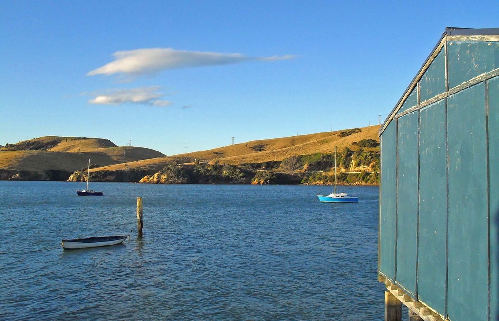 Boote und Masten im Wasser, ein hölzernes Haus rechts, mit Bäumen und Bergen mit Windmühlen im Hintergrund, unter einem bewölkten Himmel.