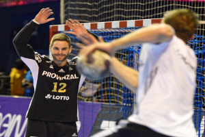 Zwei Männer spielen Handball vor einem Tor mit Netz und Banner mit Text im Hintergrund und einigen Zuschauern in einer Arena.