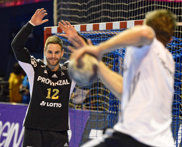 Zwei Männer spielen Handball vor einem Tor mit Netz und Banner mit Text im Hintergrund und einigen Zuschauern in einer Arena.