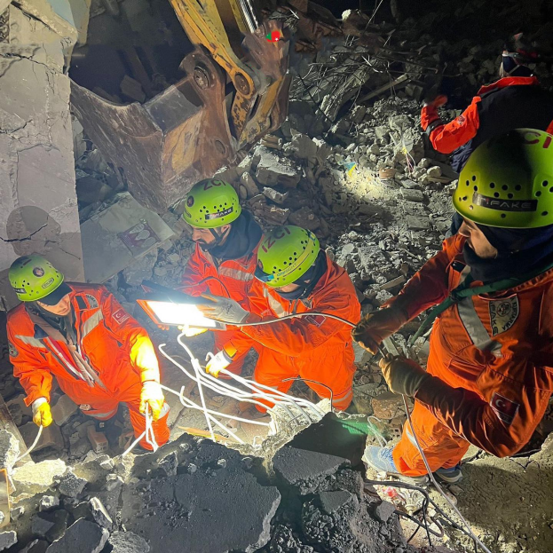 Eine Gruppe von Menschen in orangefarbenen Uniformen und grünen Helmen bei der Arbeit an Schutt mit einem Kran im Hintergrund bei einer Rettungsaktion.