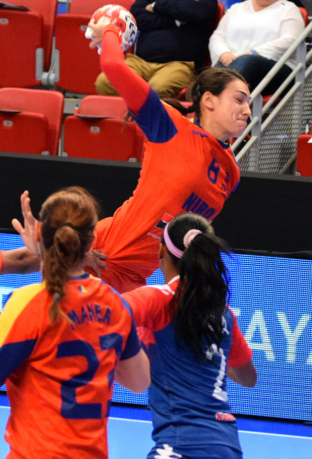 Eine Gruppe von Frauen, die Handball in einem Stadion spielen, wobei eine den Ball hält und Zuschauer im Hintergrund sitzen.