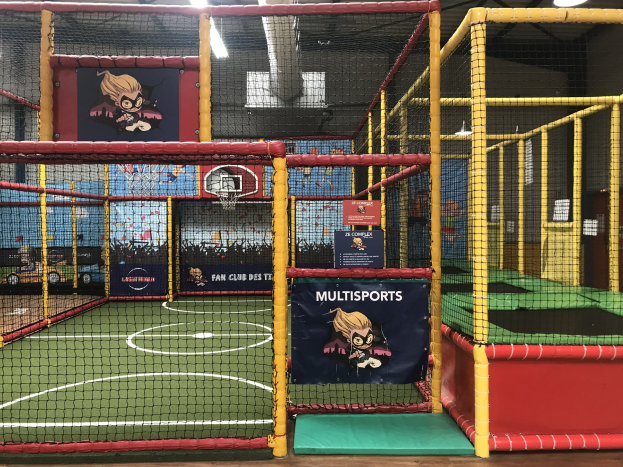 Indoor-Trampolinpark mit einem Basketballfeld in der Mitte, umgeben von einem Netz, mit einem Basketballkorb, Deckenleuchten und Wandbannern.