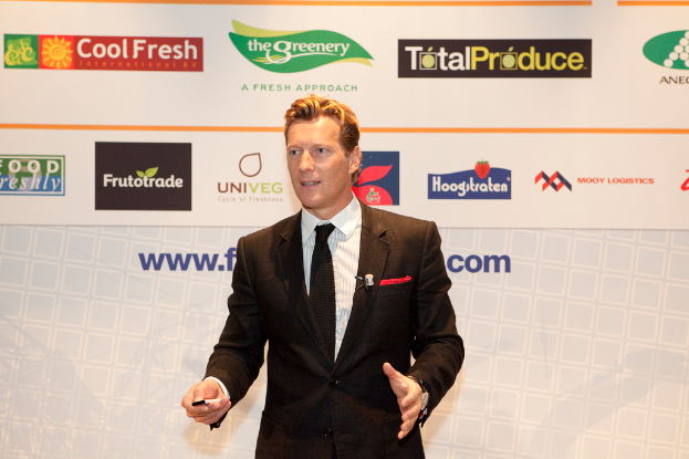 Ein Mann in einer schwarzen Blazer steht und hält ein Objekt, während er mit einer weißen Fahne und einer weißen Tafel mit organisationalen Logos im Hintergrund spricht.