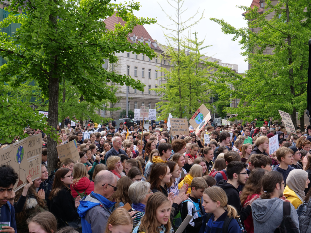 Eine große Menschenmenge protestiert vor einem Gebäude in Berlin und hält Schilder hoch, mit Bäumen, Fahrzeugen, einem Lautsprecher und Himmel im Hintergrund.