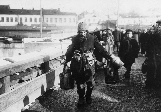 Eine Gruppe von Menschen, die wie Flüchtlinge aussehen, geht eine Straße neben einem Gewässer entlang, mit einem hölzernen Zaun links und Gebäuden und Pfählen im Hintergrund, dargestellt in Schwarz-Weiß.