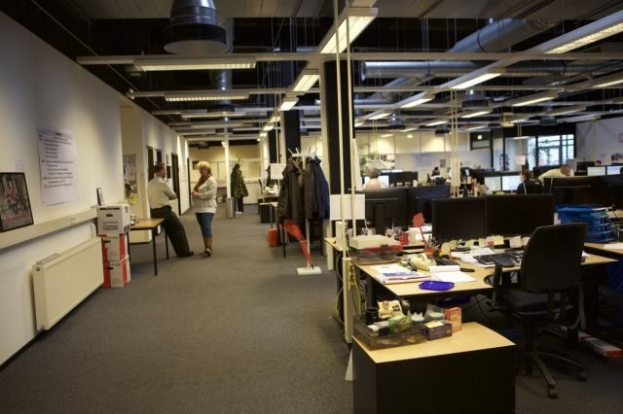 Großer open-plan-Büro mit Menschen, die an Schreibtischen arbeiten, die mit Computern, Monitoren, Tastaturen und Papieren ausgestattet sind, umgeben von Stühlen, Kisten und Büchern; Kleider hängen von Gestellen, Wände zeigen Tafeln mit Text und Bildern, Deckenlampen erhellen den Raum und Glasfenster bilden den Hintergrund.