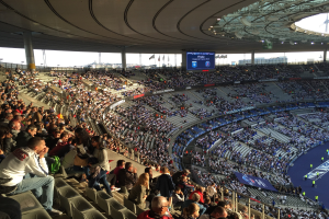 Große Menschenmenge in einem Stadion bei einem Fußballspiel, mit einer Bühne rechts, Fahnen, Stangen, einem Bildschirm und der Allianz Arena in München, Deutschland im Hintergrund.