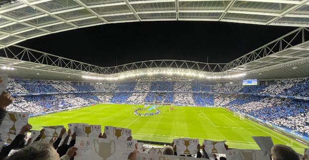 Eine Gruppe von Menschen, die Schilder in einem Fußballstadion halten, mit einigen auf dem Boden, einem Bildschirm auf der rechten Seite und Zuschauern im Hintergrund unter Stadionbeleuchtung, was eine feierliche Siegesatmosphäre vermuten lässt.