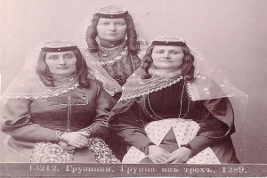 Drei Frauen in traditionellen russischen Hochzeitskleidern mit aufwendiger Stickerei und Perlenposen sich vor einer weißen Wand in einem Schwarz-Weiß-Foto, das als russisches Hochzeitsfoto bezeichnet wird.