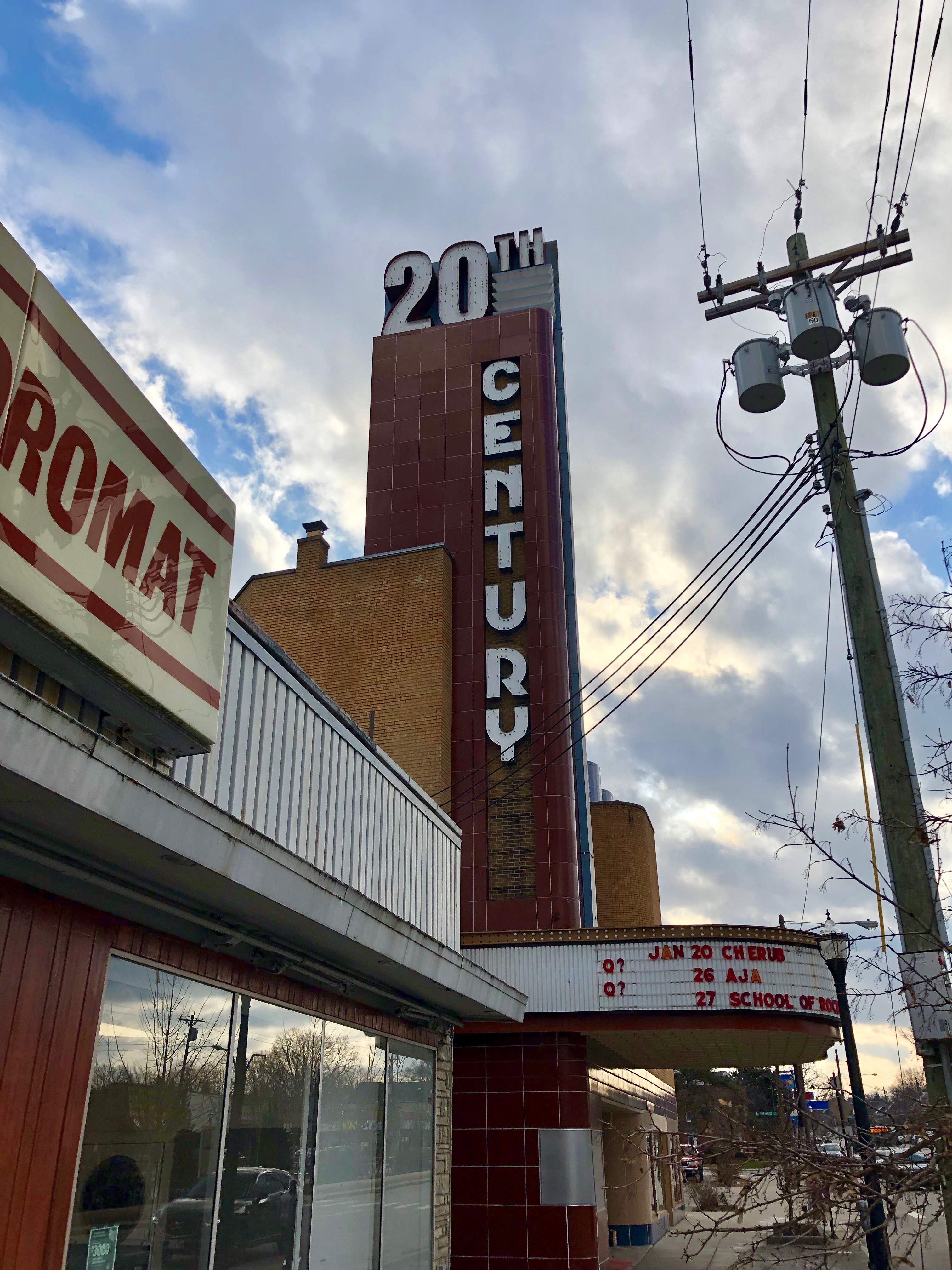 Das 20. Jahrhundert Theater in Detroit, Michigan, ein Gebäude mit Glaswänden und Namensschildern, umgeben von Bäumen und Strommasten mit Drähten, unter einem bewölkten Himmel.
