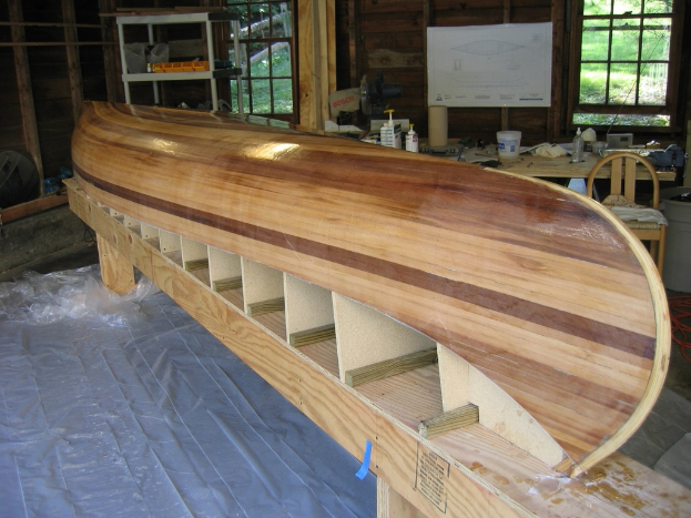 Ein Tisch mit Flaschen und Papieren, umgeben von Stühlen, mit Fenstern im Hintergrund und einer holzbootähnlichen Struktur mit Regalen dahinter auf der linken Seite.