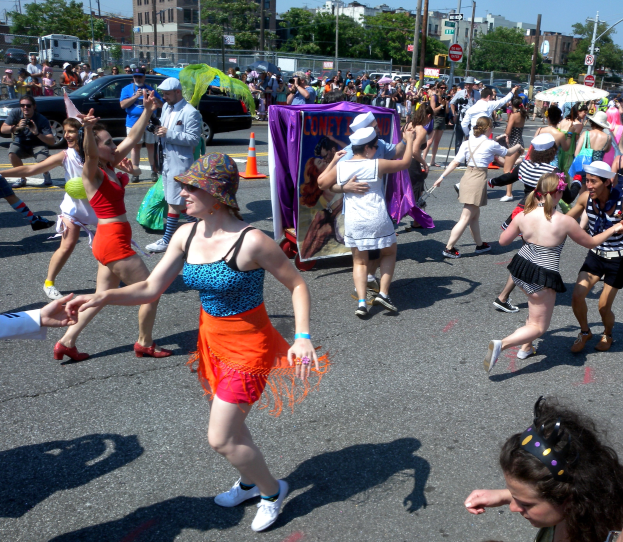 Eine Gruppe von Menschen, die auf der Straße in einer Parade gehen, einige mit Mützen und Schirmen, andere tanzen, mit einer Fahne, Verkehrskegeln, Fahrzeugen, Laternenpfählen, Schildern, Bäumen, Gebäuden und einem klaren blauen Himmel im Hintergrund.