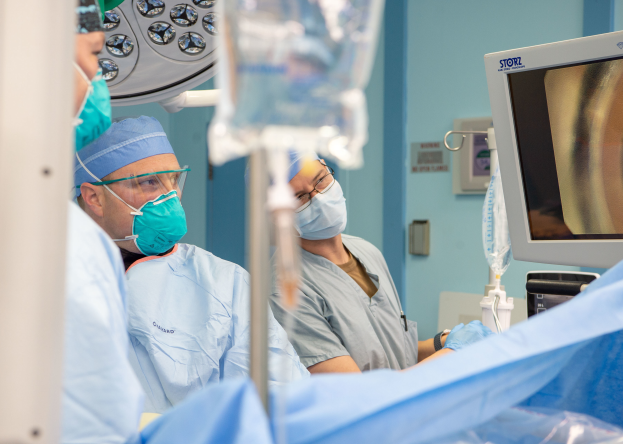 Eine Gruppe von Chirurgen in Masken, Kappen und Brillen führt einen Eingriff in einem Operationssaal durch, mit einem Monitor auf der rechten Seite und verschiedenen medizinischen Geräten, die herumliegen, mit einer Tür im Hintergrund.
