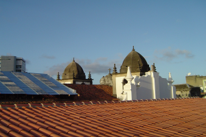 Stadtlandschaft mit Gebäuden im Vordergrund und einem blauen Himmel im Hintergrund, mit Solarpanelen auf einem Dach, die den Einsatz erneuerbarer Energien anzeigen.