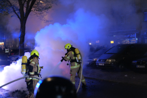 Feuerwehrleute mit Helmen und Schläuchen auf einer Straße vor einem brennenden Auto mit Rauchwolken im Hintergrund, mit Bäumen, Gebäuden und anderen Gegenständen.
