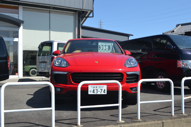 Ein roter Porsche Cayenne vor einem Gebäude geparkt, umgeben von anderen Fahrzeugen, mit Gebäuden, Polen, Drähten und einem klaren blauen Himmel im Hintergrund.