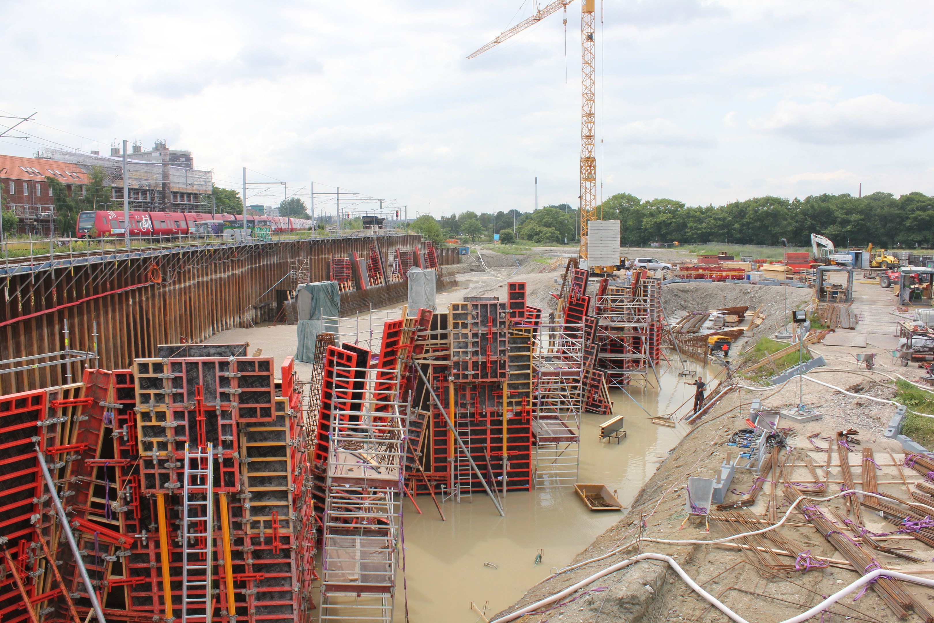 Baustelle mit Stapeln von Eisenstangen, Rohren und anderen Materialien sowie Fahrzeugen vor dem Hintergrund von Bäumen, Gebäuden, Strommasten, Drähten und einem bewölkten Himmel.