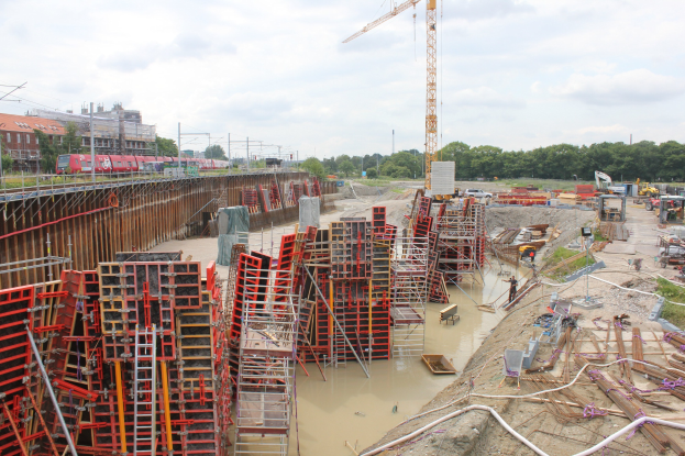Baustelle mit Stapeln von Eisenstangen, Rohren und anderen Materialien sowie Fahrzeugen vor dem Hintergrund von Bäumen, Gebäuden, Strommasten, Drähten und einem bewölkten Himmel.