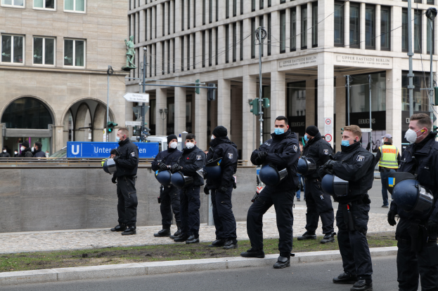 Eine Gruppe von Polizisten in schwarzen Uniformen und Masken steht vor einem Gebäude mit Glasfenstern und Säulen in Berlin, Deutschland, einige halten Helme, mit Laternen, Verkehrsampeln, Schildern mit Text, einer Statue, anderen Menschen und einer grasbewachsenen Fläche im Hintergrund.